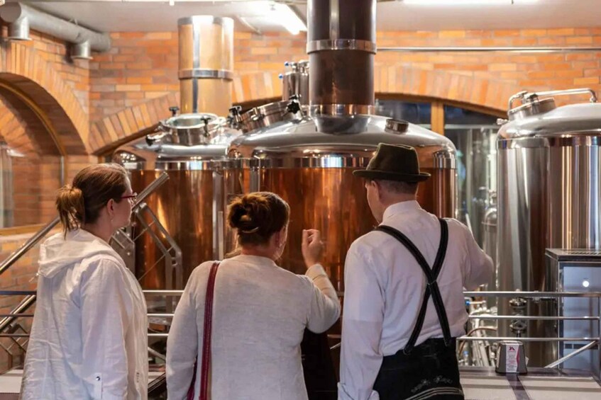 Three people observing industrial brewing equipment in a brick-walled facility. 1