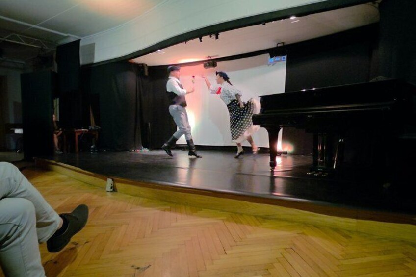 A stage with a piano, a man and a woman performing, and a large screen in the background. 3