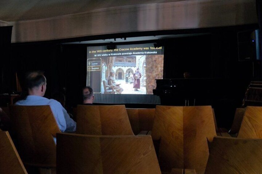 A lecture hall with wooden chairs, a screen displaying a presentation, and a person sitting in the audience. 4