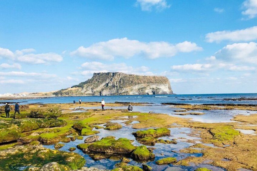 A rocky shoreline with people walking, a large cliff in the background, and a clear sky. 2
