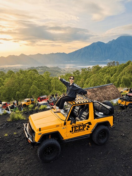 A person on a yellow Jeep with 'BATUR INSIDE JEEP ADVENTURE' text, surrounded by vehicles and people in a mountainous area. 2
