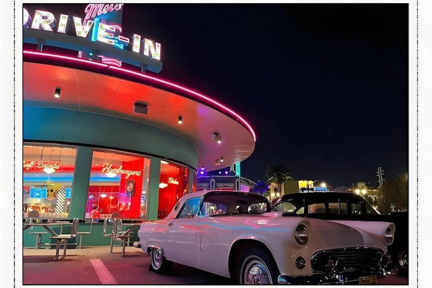 A vintage car in front of a neon-lit drive-in theater with signs for hamburgers and hot dogs. 5