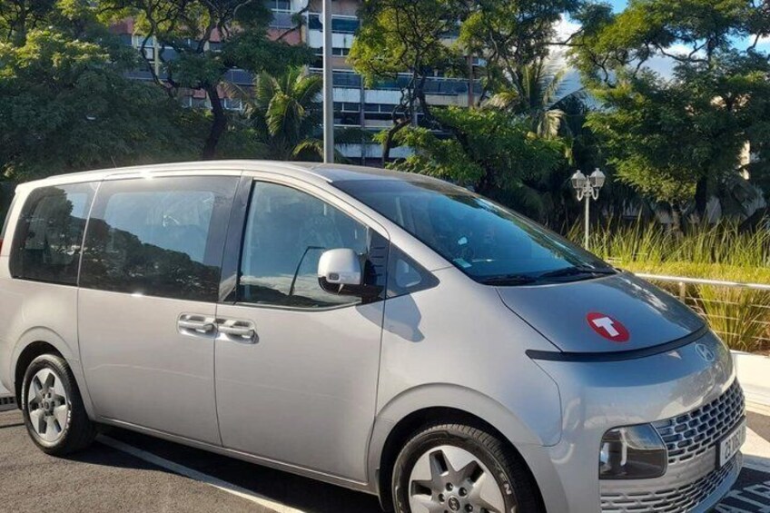 A silver minivan parked on a street with trees and buildings in the background. 3