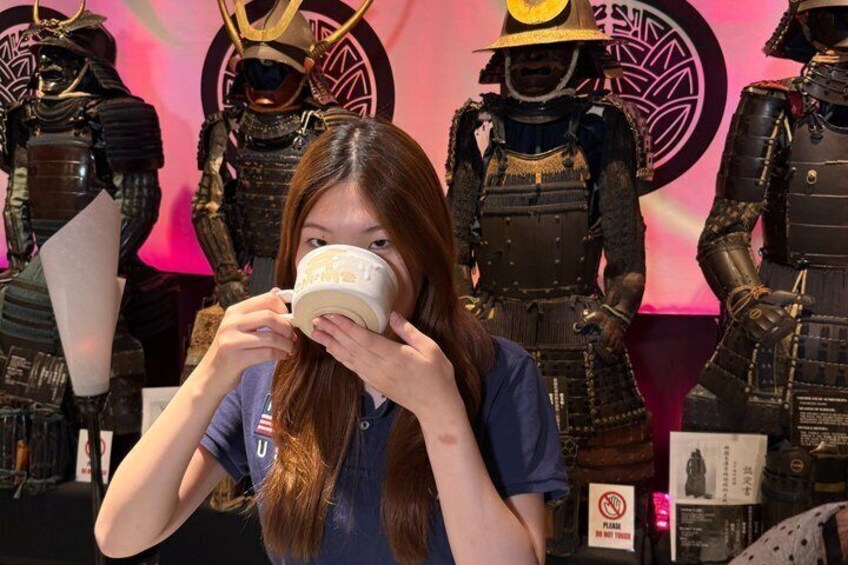 A person holding a mug in front of a display of samurai armor. 3