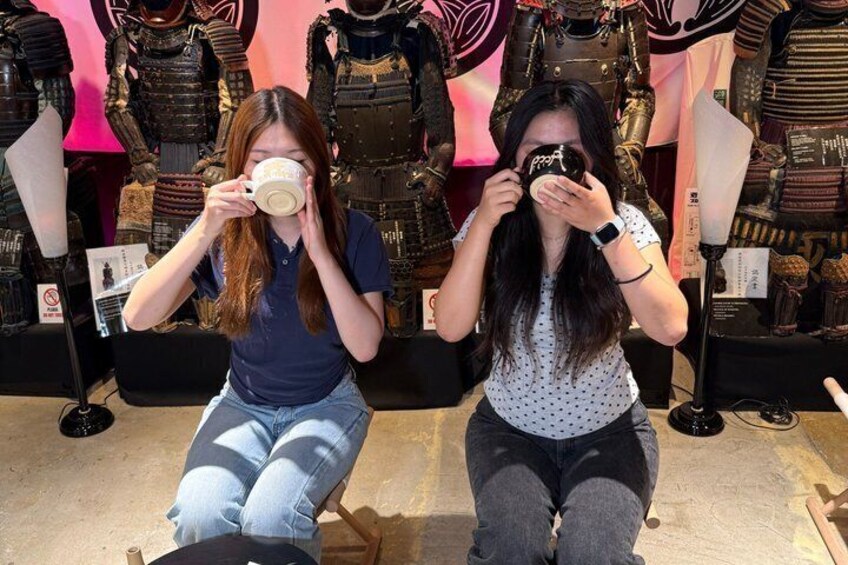 Two individuals seated in front of a display of armor and protective gear. 2