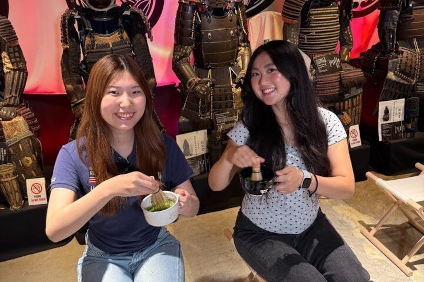 Two people sitting at a table with food, in a room with various items on display. 1