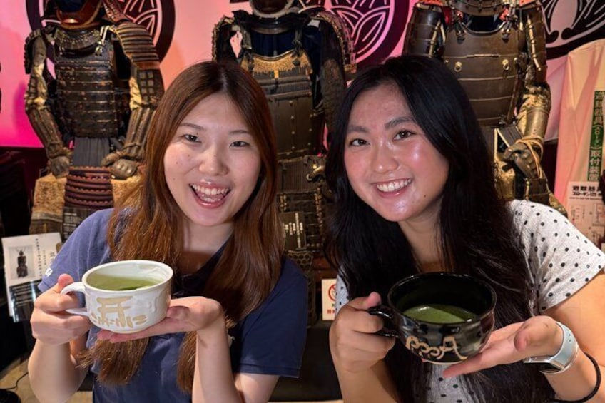 Two individuals holding cups, with a display of armor in the background. 5