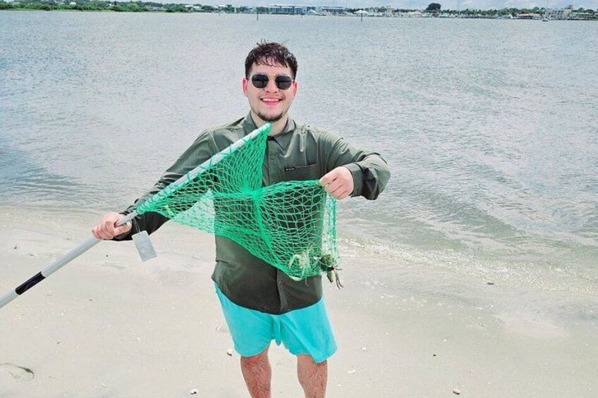 A person holding a green fishing net on a sandy beach with water in the background. 2