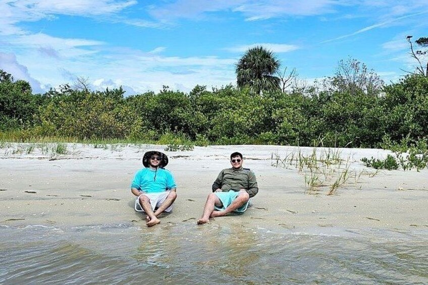 Two people sitting on a sandy beach with water around them and vegetation in the background. 3