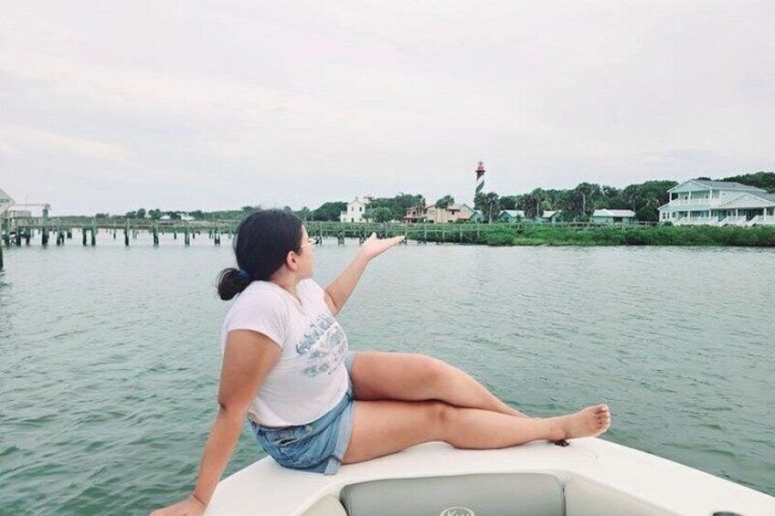 A person sitting on a boat, facing a body of water with a lighthouse and buildings in the background. 2