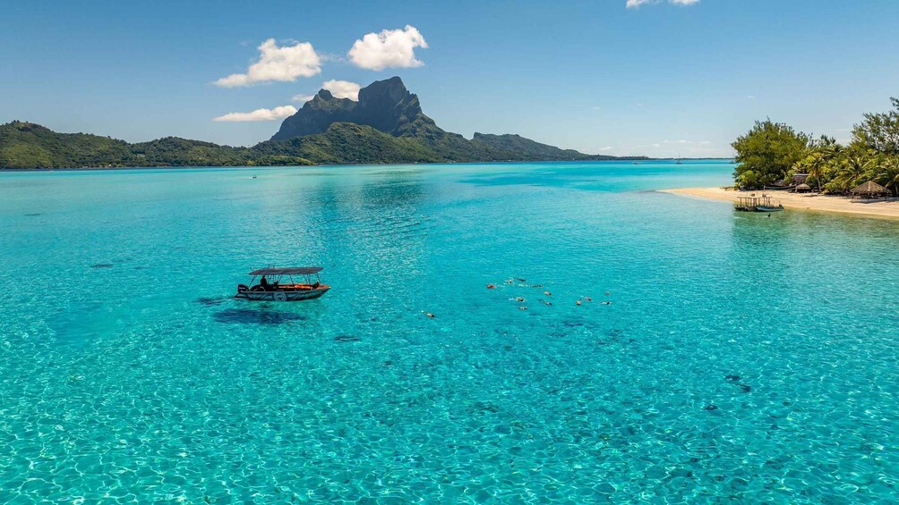 A clear turquoise sea with a boat and snorkelers, a sandy beach, and a mountain in the background. 5