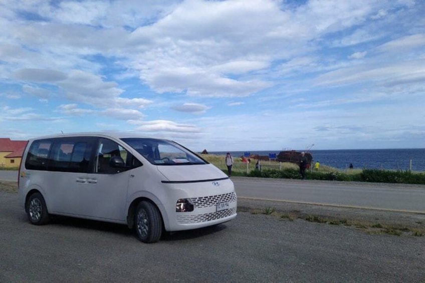 A white minivan parked on a road with a beach and ocean in the background. 1