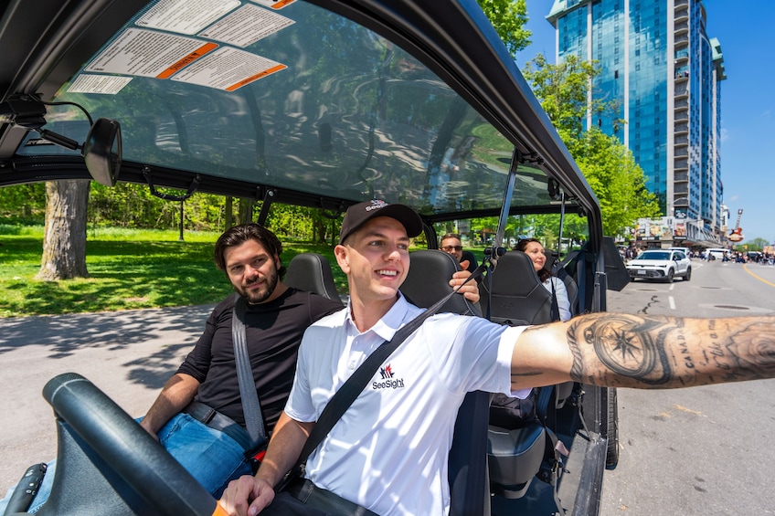 Une personne se trouve dans un véhicule de golf, son bras est tatoué, elle porte une chemise blanche ornée d’un logo, et une autre personne est à ses côtés, vêtue d’une chemise noire. 3