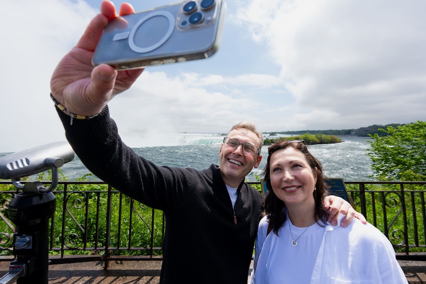 Une personne prend un autoportrait avec un smartphone, une autre personne se tient à côté, un télescope et une rivière avec une végétation luxuriante en arrière-plan. 4