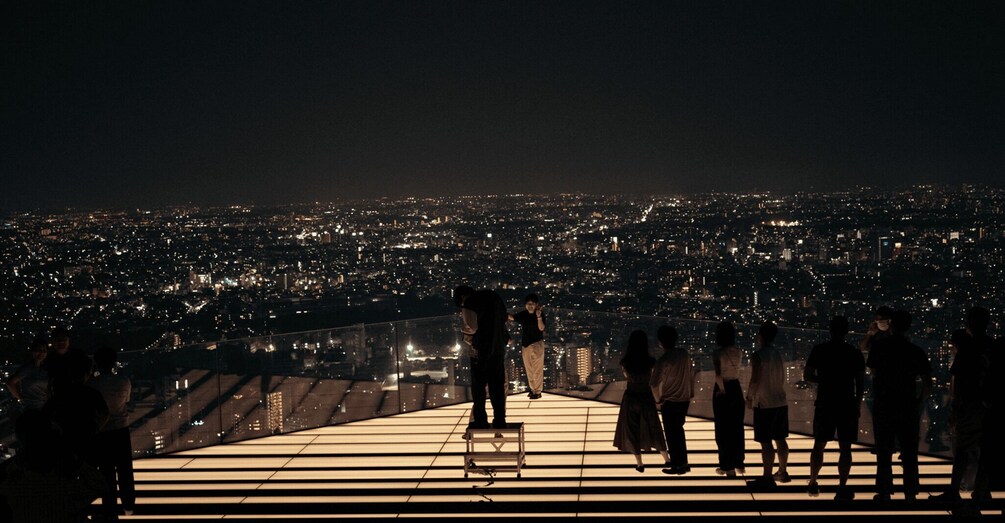 A group of people standing on a balcony overlooking a cityscape at night. 1