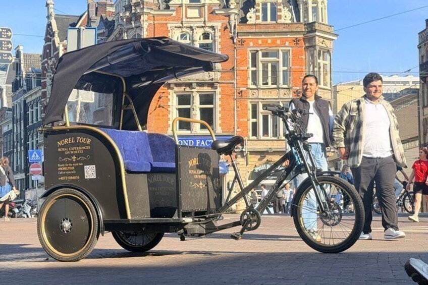 A vintage bicycle carriage with a black canopy, a man standing beside it, and a bicycle leaning against it. 3