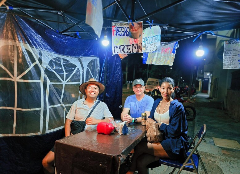 A group of people sitting at a table under a tent with signs advertising food and drinks. 2