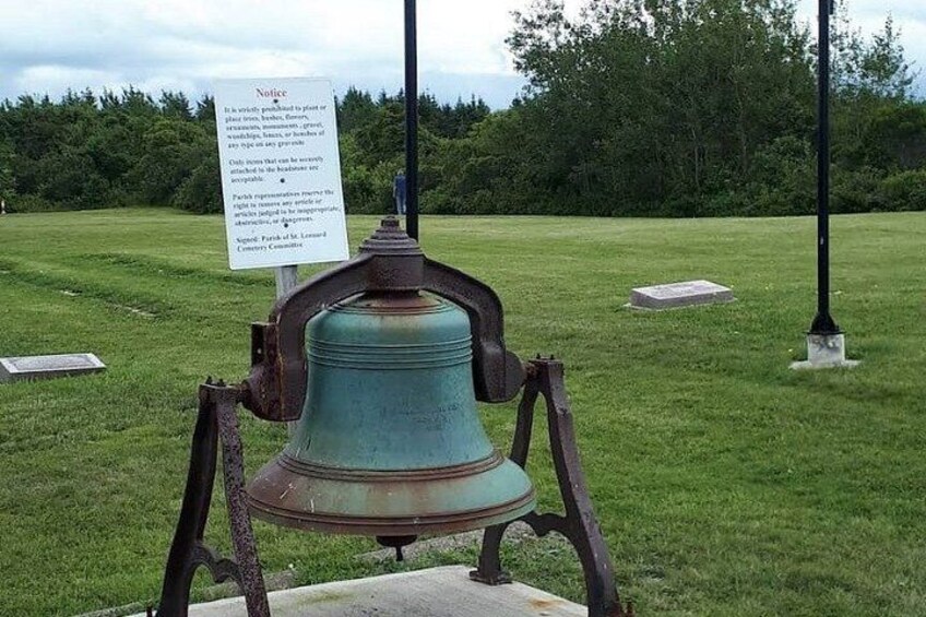 A large, weathered bell mounted on a metal stand in a grassy field with a sign in the background. 1