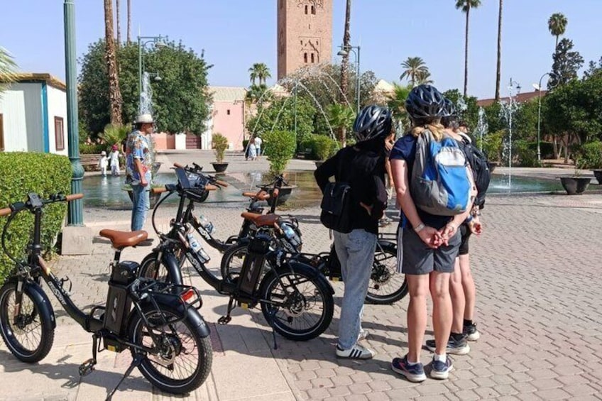 A group of people standing near bicycles in a paved area with a fountain and palm trees in the background. 5