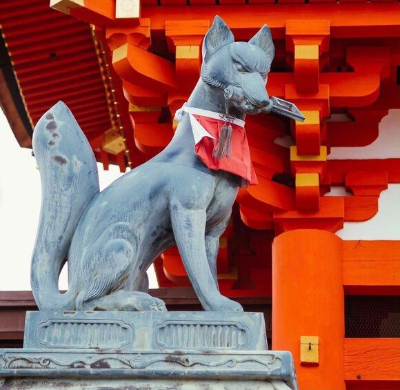 Punaisella kaulapannalla ja kellolla varustetun ketun patsas, jonka taustalla on punavalkoinen, perinteinen japanilainen rakennus. 3