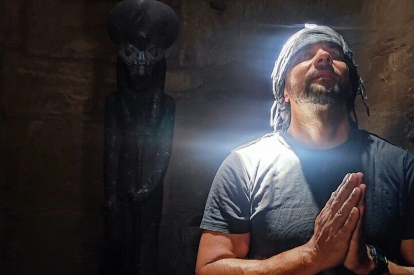 A person in a gray shirt with hands clasped in prayer, standing in front of a dark, textured wall. 1