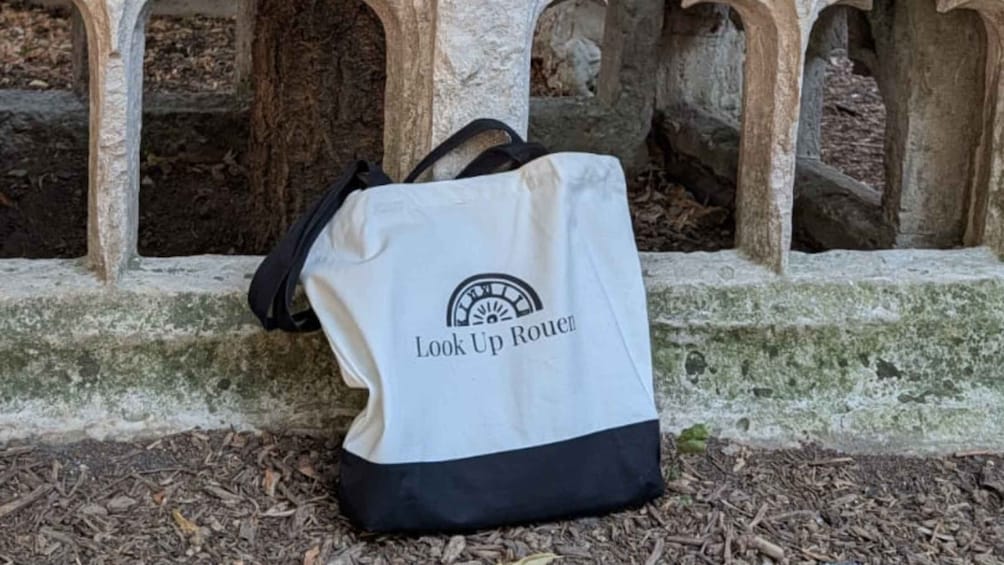 A white and black tote bag with the text 'Look Up Rouen' on it, placed on a stone ledge with moss. 2