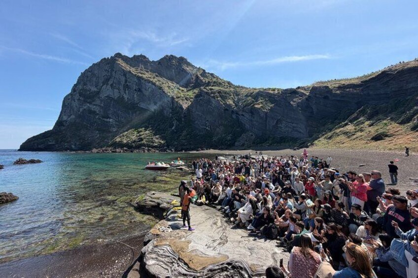 A rocky shoreline with a crowd of people gathered, a clear blue sky, and a mountain in the background. 5