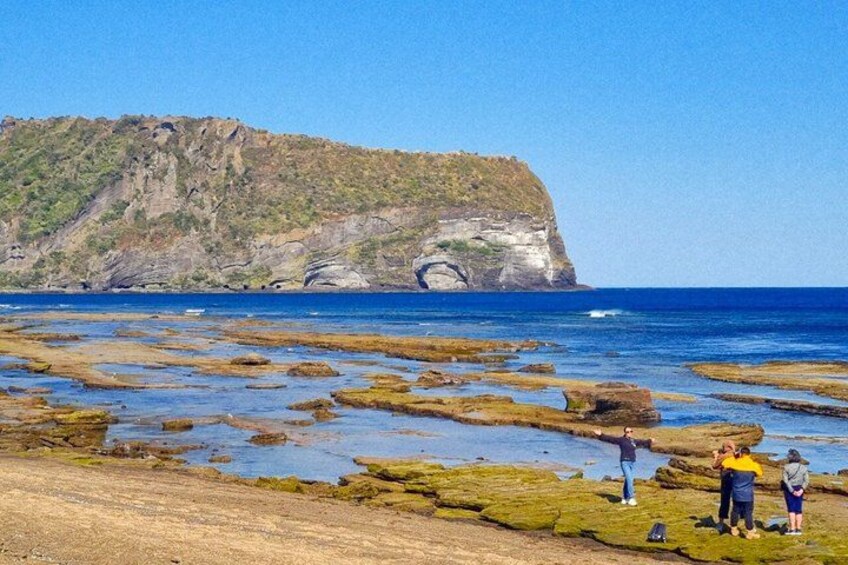 A rocky shoreline with people standing on the beach, a cliff in the background, and calm blue water. 2