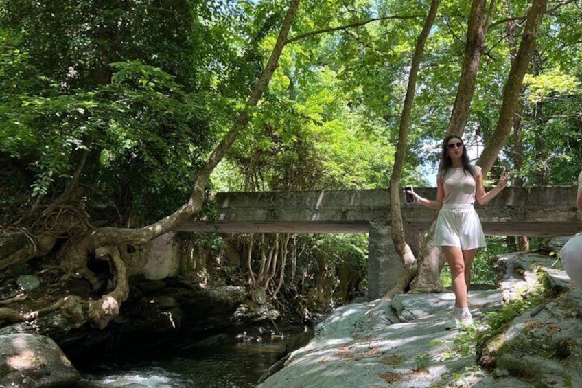 A woman in a white dress standing on a rock by a stream in a forested area. 1