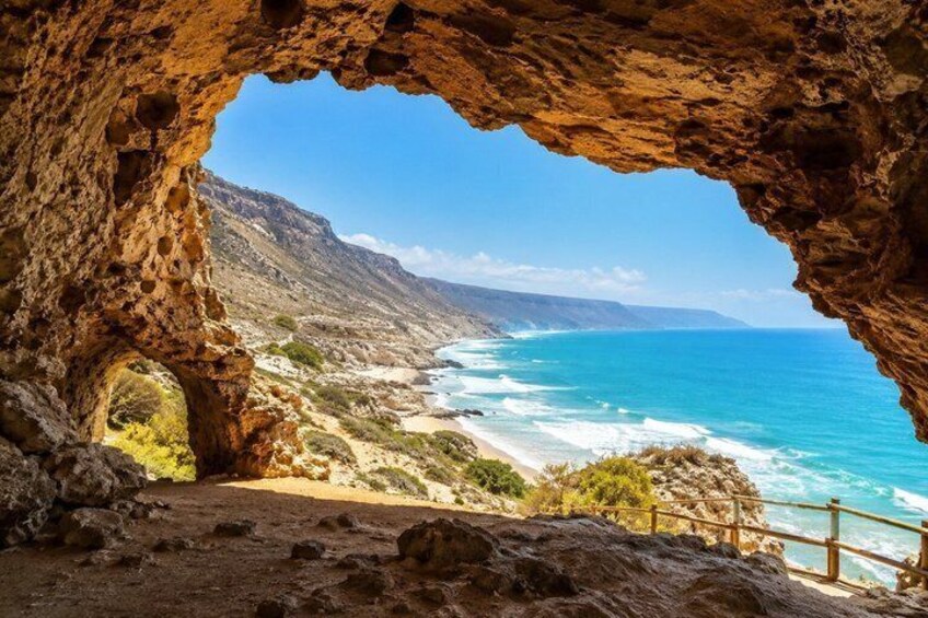A coastal landscape with a rocky cave entrance, a sandy beach, and the ocean. 1