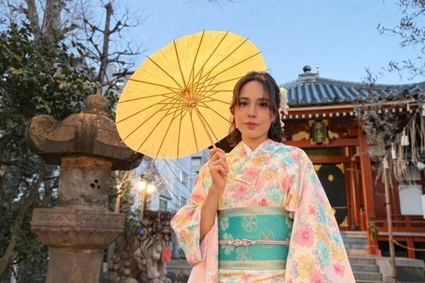 A person in traditional Japanese attire holding a yellow fan, standing in front of a stone lantern and a traditional building. 3