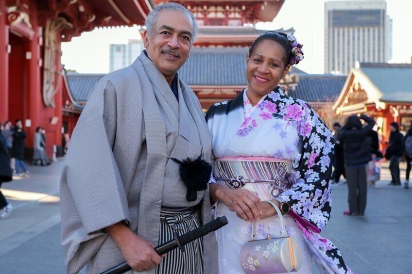 A person in traditional Japanese attire standing in front of a red building with a modern cityscape in the background. 4