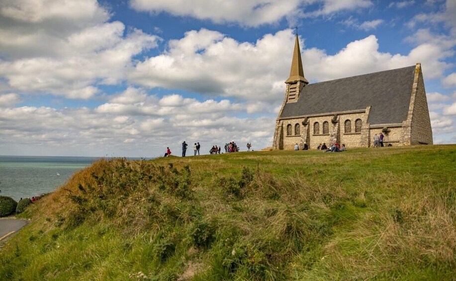 A stone church with a tall steeple, a grassy hill, and a body of water in the background. 3
