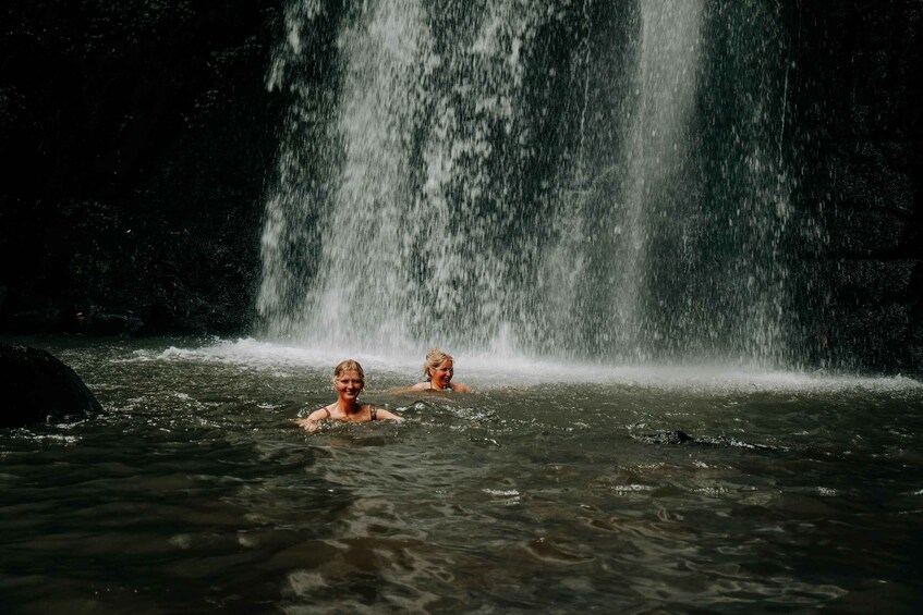 Two people swimming in a waterfall pool with a dense forest in the background. 3