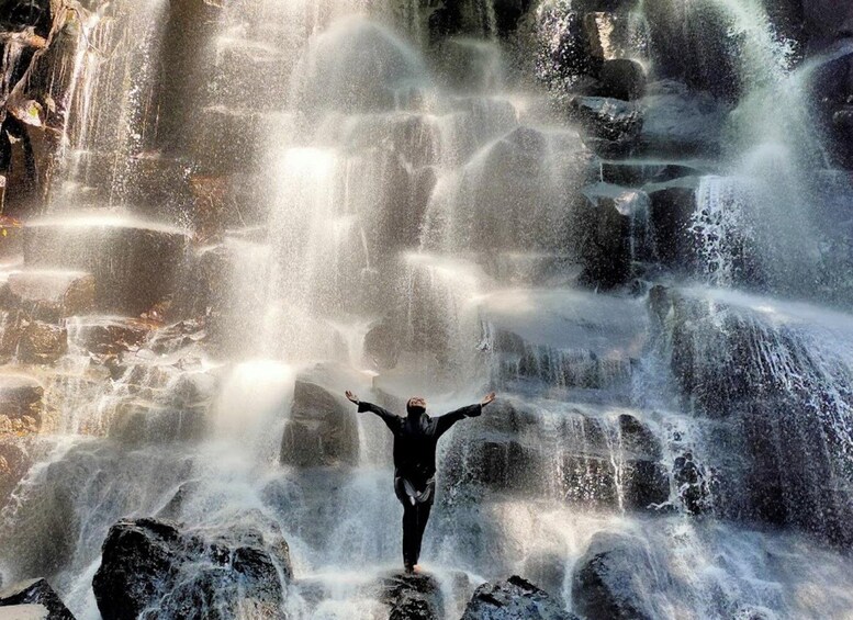 A person standing on rocks with arms outstretched in front of a waterfall. 1
