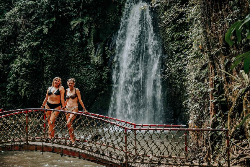 Two people on a rope bridge over a waterfall in a forested area. 2