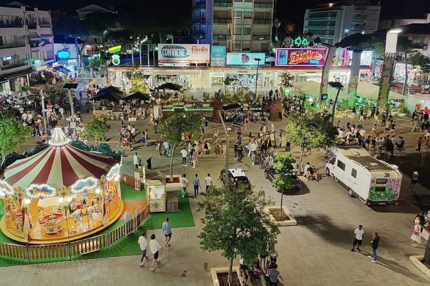 A crowded urban square with a carousel, food stalls, and a large RV parked in the center. 2