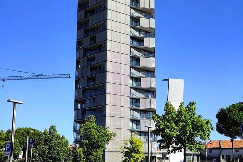 A tall residential building with multiple balconies, a construction crane in the background, and trees in the foreground. 3