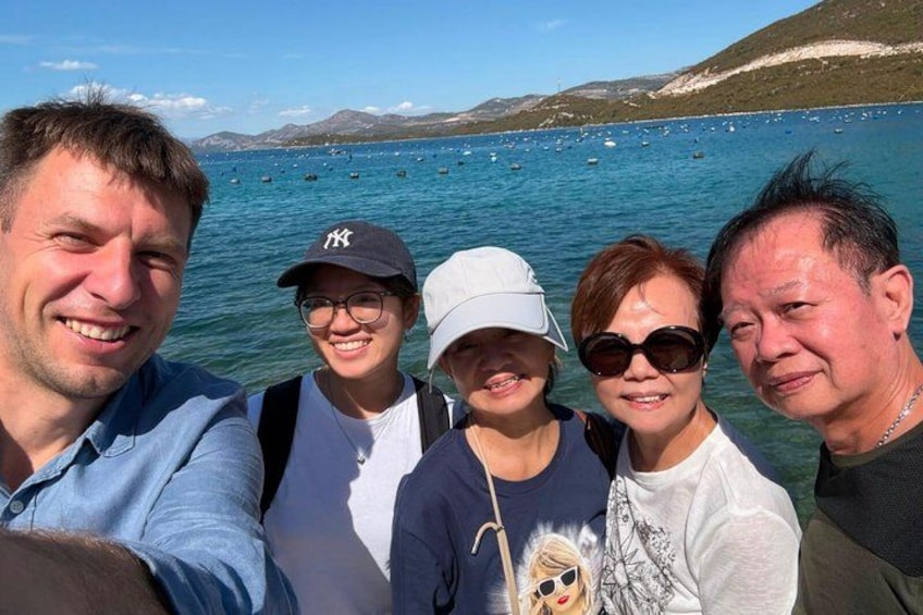 A group of people taking a selfie by a body of water with boats and hills in the background. 3