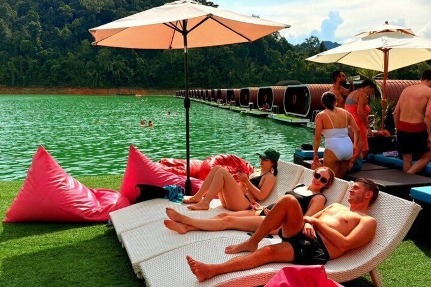 People relaxing on lounge chairs by a lake with inflatable pink pillows and umbrellas. 1