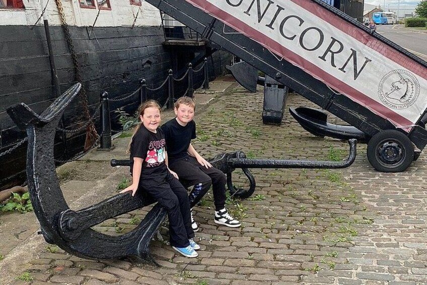 Two people sitting on a large anchor sculpture with a banner reading 'UNICORN' in a cobblestone area. 3