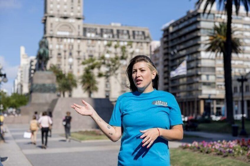 A person in a blue shirt with a logo, standing in an urban setting with buildings and palm trees in the background. 1