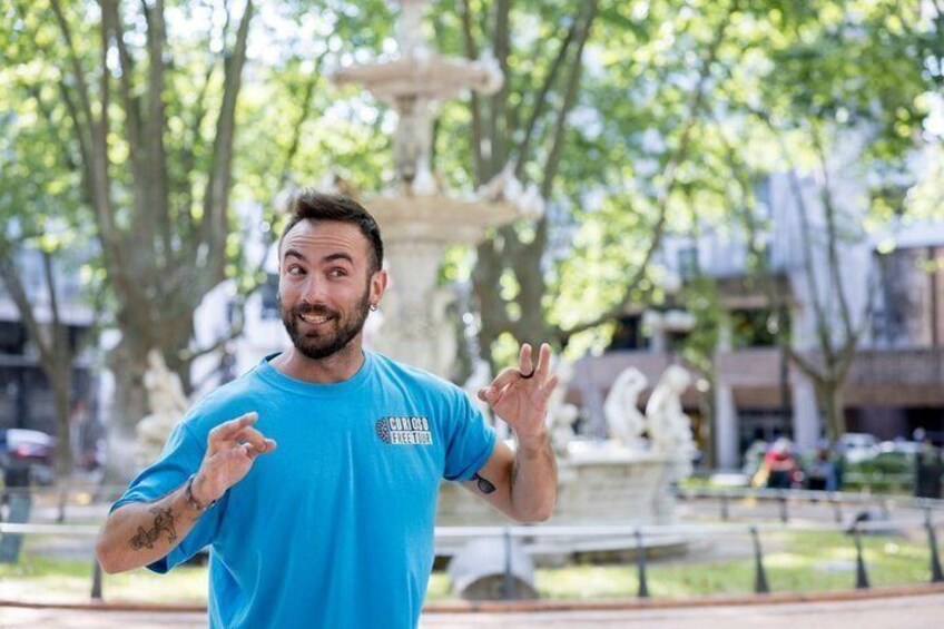 A person in a blue shirt with a logo, standing in a park with a fountain and trees in the background. 2