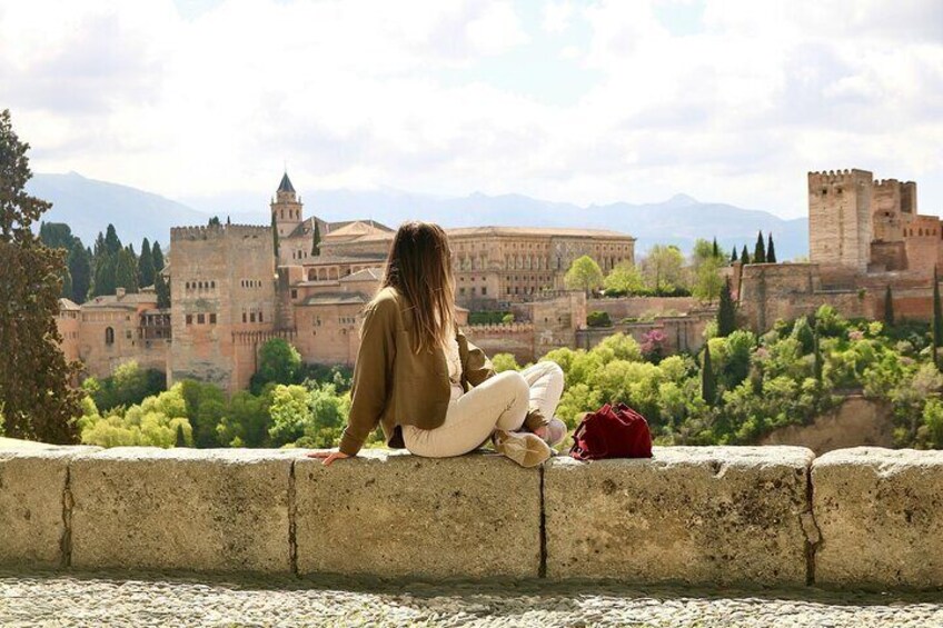 A person sitting on a stone wall overlooking a historic city with ancient buildings and greenery. 1