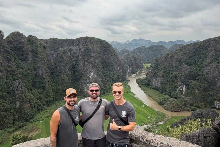 Three people standing on a cliff overlooking a river valley with dense greenery and rocky terrain. 1