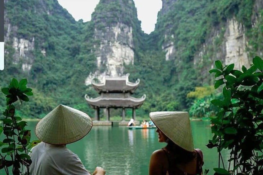 Two people in traditional conical hats overlooking a temple on a river surrounded by cliffs. 4