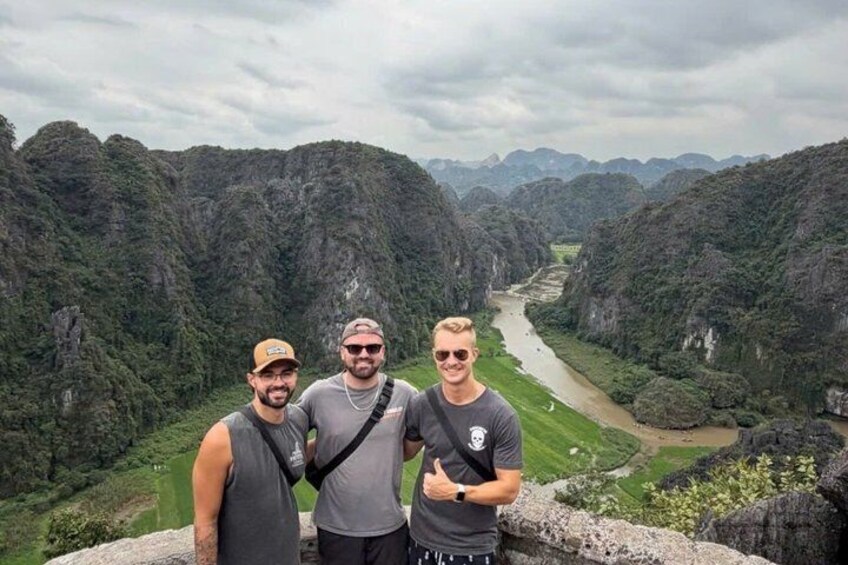 Three people standing on a cliff overlooking a river valley with dense vegetation and rocky terrain. 1