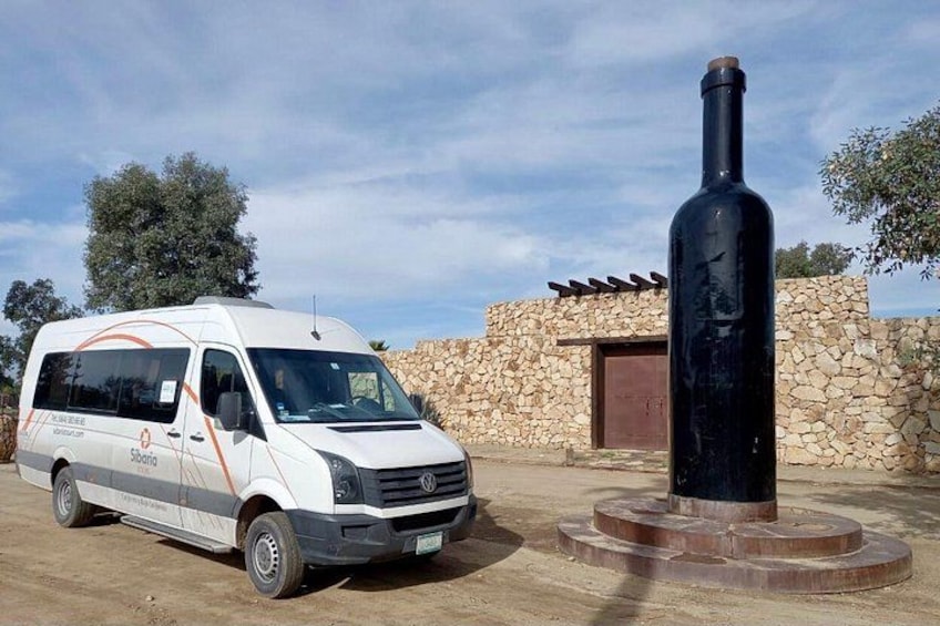 A white van parked in front of a stone wall with a large black bottle sculpture. 2