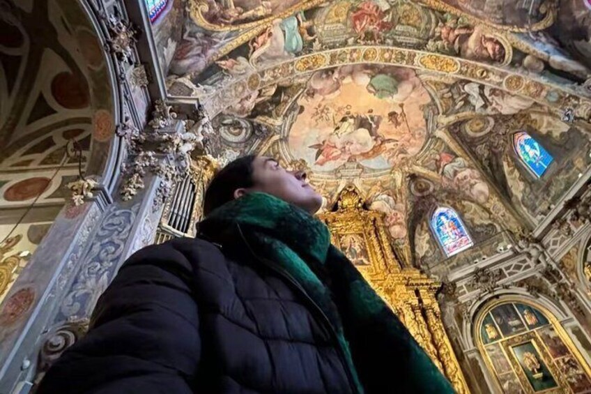 A person taking a selfie inside a church with ornate ceiling artwork and stained glass windows. 1
