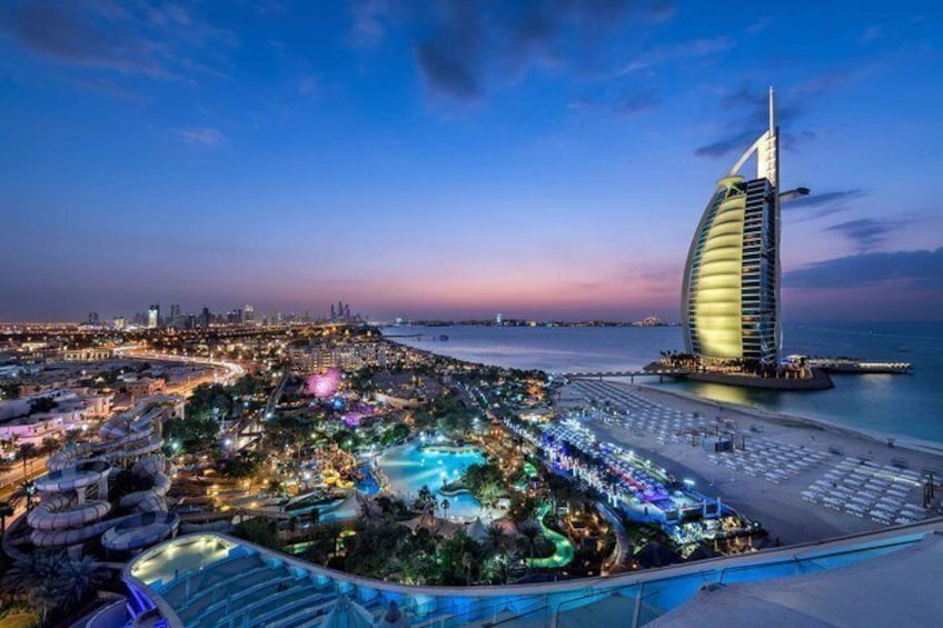 A coastal cityscape at dusk with a prominent sail-shaped building, a well-lit beachfront, and a vibrant entertainment complex. 1